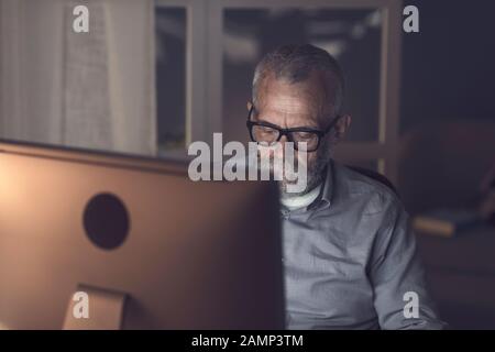 Senior man sitting at desk and working with his computer late at night Stock Photo