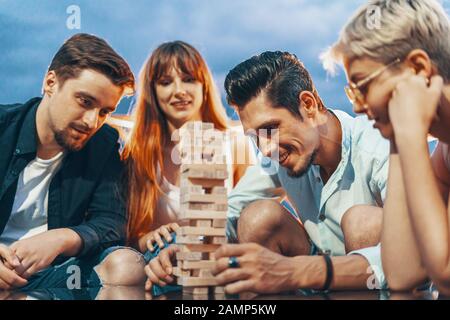 The company of young people playing jenga. Young friends at sunset on penthouse patio playing jenga Stock Photo