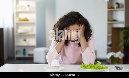 Young biracial woman choosing between salad and sweets, health care choice Stock Photo