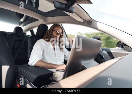 Mode of Transport. Bearded man with long hair in sunglasses traveling sitting inside electric car driving on autopilot browsing laptop joyful Stock Photo