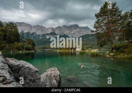 Woman swimming in the lake, rocks on the shore, view into the distance, Eibsee lake in front of Zugspitze massif with Zugspitze, Wetterstein range Stock Photo