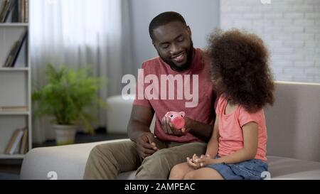 Smiling father helping daughter save money in piggy bank for presents, finance Stock Photo