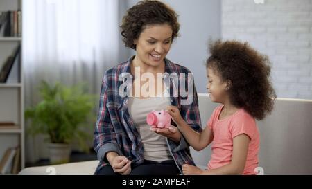 Mother teaching little daughter to save money, throwing coins in piggy bank Stock Photo