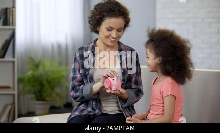 Mom and daughter throwing coins into piggy bank, saving money for presents Stock Photo