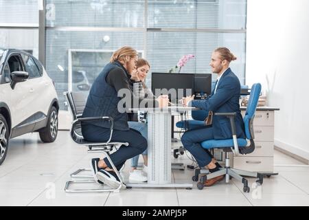 Car Salon. Young couple sitting at table with agent buying car reading contract joyful side view Stock Photo