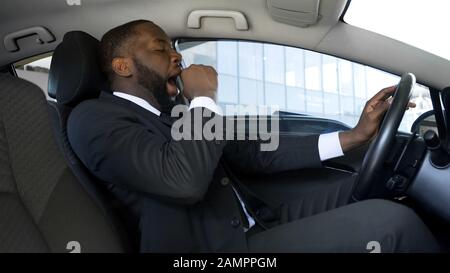 Tired black man yawning in car, overworked businessman driving car, danger Stock Photo
