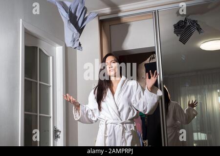 General plan Portrait of a young beautiful girl in a home white coat, sitting on the floor in a room with scattered things, tossing clothes. In the ea Stock Photo