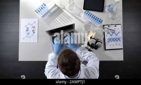 Lab worker viewing samples under microscope and typing results on laptop topview Stock Photo