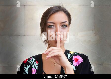 Close-up portrait of a beautiful brunette girl holding a finger on her lips asking for silence, shh! Stock Photo