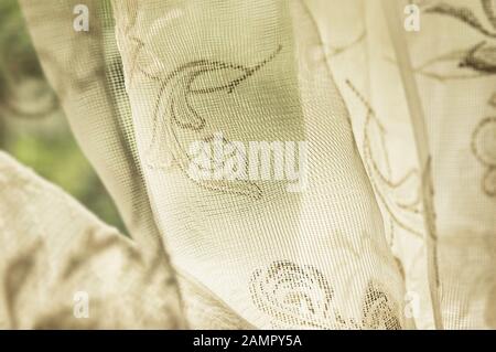Close up detail of old fashioned sheer fabric patterned curtain hanging on window lit with natural light Stock Photo