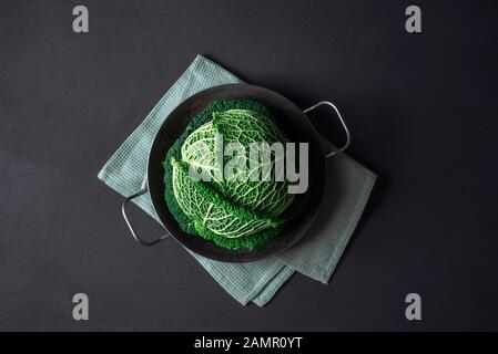 Green cabbage in a rusty iron pan on a kitchen towel on black background. Healthy eating. Diet food. Overhead of savoy cabbage. Fresh vegetables. Stock Photo