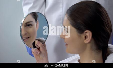 Young female satisfied with rhinoplasty result, smiling face reflected in mirror Stock Photo