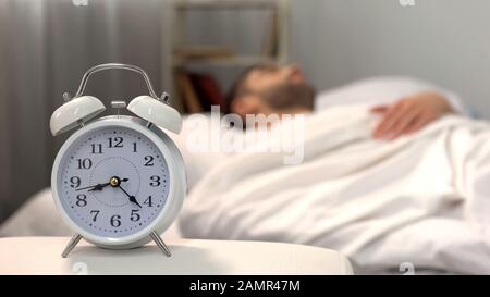 Male sleeping in bed, white alarm clock standing on table morning, biorhythm Stock Photo