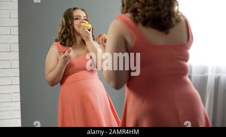 Happy obese lady satisfied with her body eating sweet donuts in front of mirror Stock Photo