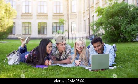 Multi-ethnic students lying on grass and watching funny video on laptop, friends Stock Photo