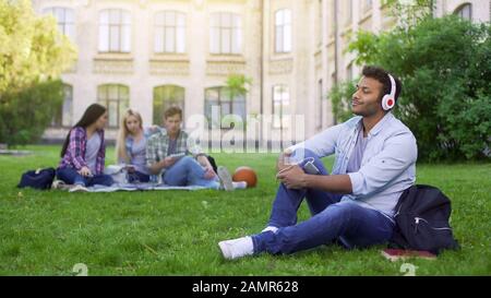 Handsome hispanic student sitting on grass and enjoying music in headphones Stock Photo