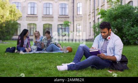 Serious biracial male student sitting on grass and writing essay, doing homework Stock Photo