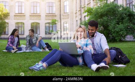 Multi-ethnic students sitting on grass and watching funny video on laptop Stock Photo