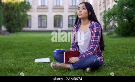 Thoughtful asian female student sitting on lawn alone and missing parents Stock Photo