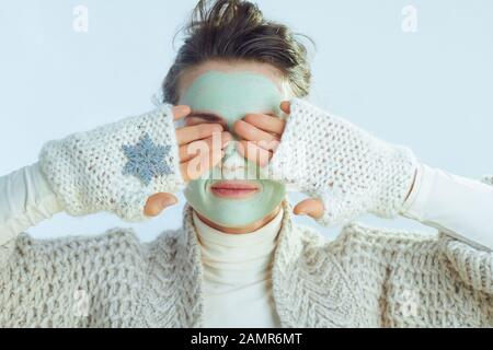 modern housewife in roll neck sweater and cardigan with green facial mask, eyes covered with hands showing snowflake isolated on winter light blue bac Stock Photo