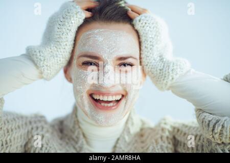 Portrait of cheerful elegant middle age housewife in roll neck sweater and cardigan with white facial mask isolated on winter light blue background. Stock Photo