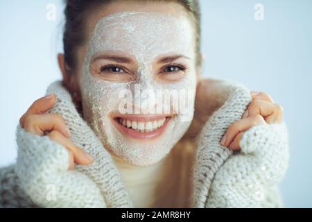 smiling stylish woman in roll neck sweater and cardigan with white facial mask covering in clothes on winter light blue background. Stock Photo