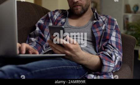 Man sitting on sofa and typing on laptop, scrolling smartphone, applying for job Stock Photo