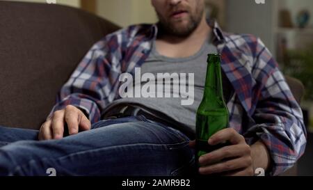 Depressed drunk man sitting on couch and drinking bottled beer, money problems Stock Photo