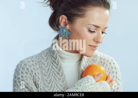 elegant woman in roll neck sweater and cardigan holding orange isolated on winter light blue background. Stock Photo