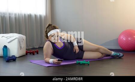 Lazy fat female lying on mat scrolling social media on gadget instead of workout Stock Photo