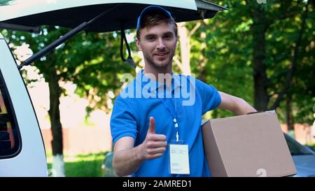 Young delivery man holding parcel, showing thumbs up, fast shipping service Stock Photo