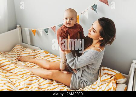 A little baby girl smiles and plays on the bed with her mom, dressed in knitted sweaters pastel fashionable shades, in a room, cozy house, modern design. Happy Family at home, concept of motherhood Stock Photo