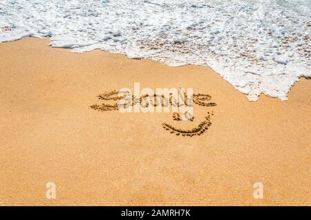 Hand written text smile and symbol on the golden beach sand with coming wave. Cheer up and fun concept. Summer holidays and vacation at the sea. Relax Stock Photo