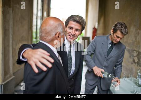 Mid-adult business man talking to a senior male colleague has an arm around his shoulder while a mid-adult co-worker is pouring water in a courtyard. Stock Photo