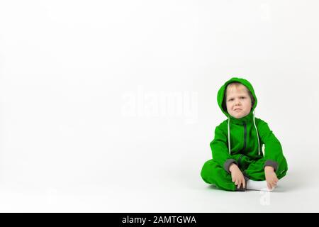 A displeased charismatic child in a bright green overalls sits on the floor and looks at the camera with doubt and contempt. Concept on a white backgr Stock Photo