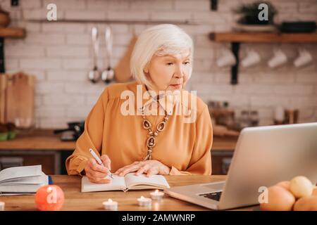 Grey-haired beautiful elderly lady in orange blouse reading a lesson online Stock Photo