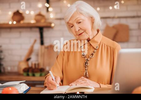 Grey-haired beautiful elderly lady in orange blouse making notes Stock Photo