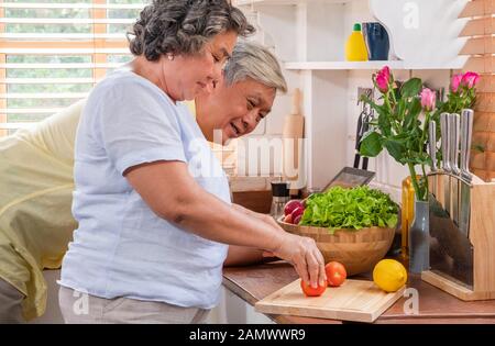 Asian senior wife slicing tomatoes on wooden cutting  board and using tablet computer to searching menu recipe.senior with technology lfiestyle.aging Stock Photo