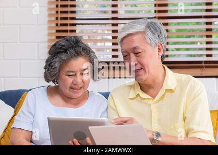 Asian senior couple use tablet searching about retirement financial document sitting on sofa at home,senior learn to use technology.aging in place con Stock Photo