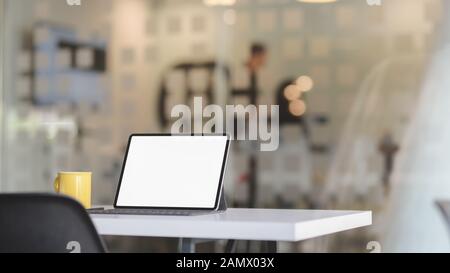 Cropped shot of simple workplace with blank screen tablet and coffee cup on white table with blurred  office room background Stock Photo