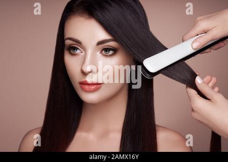 Hairdresser straightening long dark hair with hair irons. Beautiful woman with long straight hair. Smooth hairstyle Stock Photo