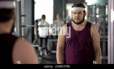 Fat man looking at mirror reflection in gym, mental training before workout Stock Photo