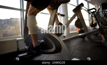 Athlete male with bandaged leg riding stationary bike in morning, recovery Stock Photo