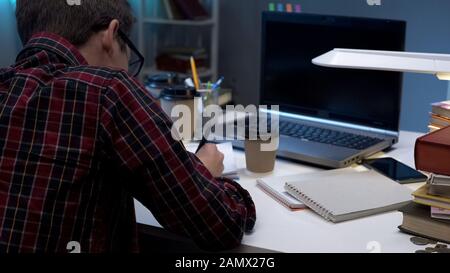 Teen male in reading glasses writing essay overworked student preparing for exam Stock Photo