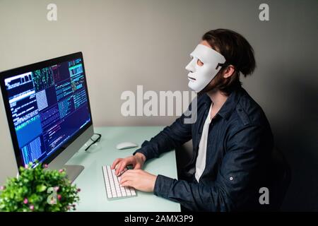 Anonymous computer hacker in white mask and hoodie. Obscured dark face making silence gesture in Underground Secret Location Surrounded by Displays an Stock Photo