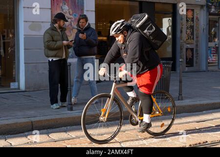 Milan, Italy - January 11, 2020: A courier on bike deliver tasty food in a city centre street. Food delivery service. Online ordering. Stock Photo