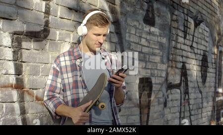 Young man listening to music and scrolling on cellphone, holding skateboard Stock Photo