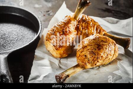 Three spicy seasoned grilled chicken legs on crumpled white paper alongside an old vintage metal skillet Stock Photo