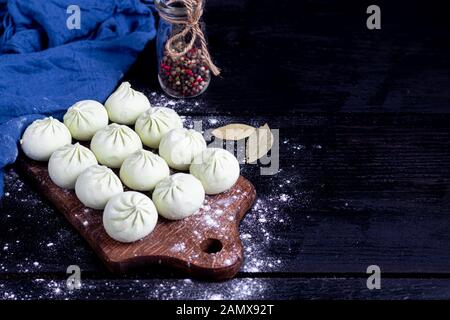 Frozen khinkali on a wooden board, sprinkled with flour. In the background pepper and bay leaf. On a black wooden background. Copy space. Stock Photo