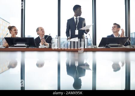 African young businessman standing and performing with financial report and people clapping to him at conference Stock Photo